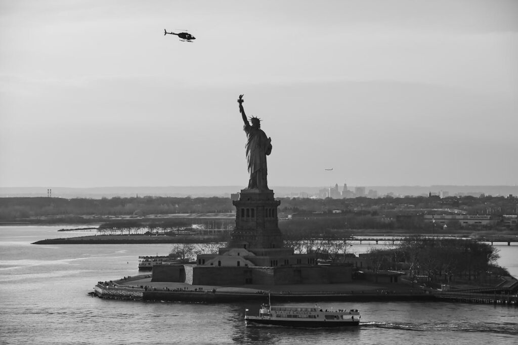 Black and white aerial photograph of the Statue of Liberty with a helicopter overhead and a ferry below.